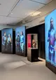 Drei menschhohe Monitore, auf denen Personen zu sehen sind, in der Mitte eine Vitrine  mit einer Figur darin.