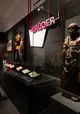 Dunkler Raum mit mehreren Schriftstücken in einer Vitrine, links ein Monitor mit einer Person und rechts eine Holzstatue.