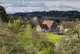Landschaft mit Büschen und Bäumen im Vordergrund, mittig ein Haus mit großer Scheune und im Hintergrund Wälder und Berge.